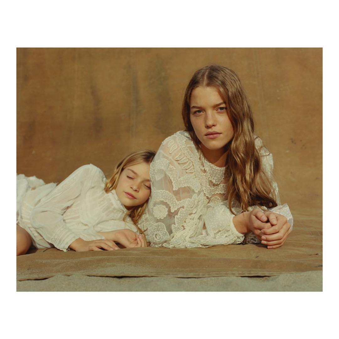Two young girls in white dresses pose in a studio setting with a neutral-toned backdrop and soft lighting, showcasing their natural beauty.