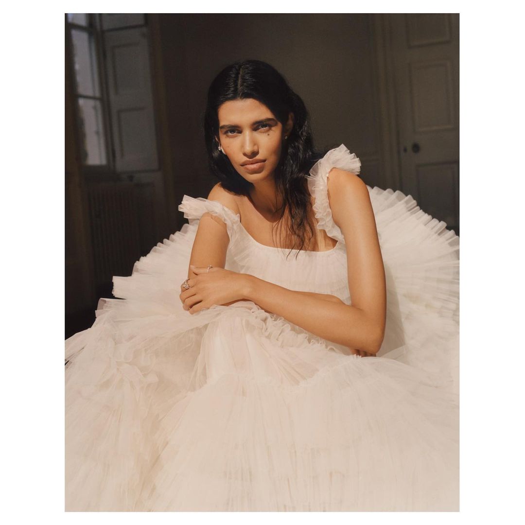 A woman in a white tulle gown with ruffle straps poses indoors. The lighting is warm and casts shadows in the background.