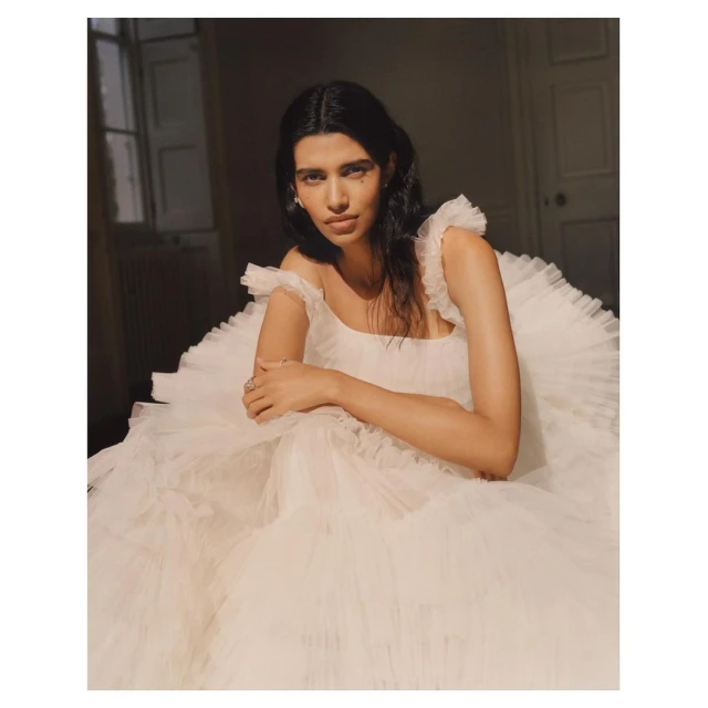 A woman in a white tulle gown with ruffle straps poses indoors. The lighting is warm and casts shadows in the background.