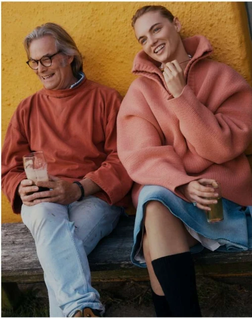 Couple sits on wooden bench outside. Man in glasses holds drink. Woman in pink sweater, jean skirt and boots. Yellow background.