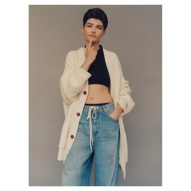 Woman in casual attire, wearing a cardigan and jeans, stands in a studio with hand to face in a thinking pose, in a medium shot.