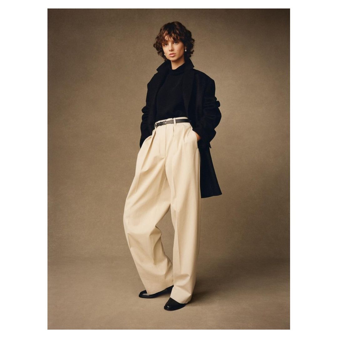 Studio shot of an adult woman wearing a black long-sleeved shirt, oversized cream pants, and a black coat, standing against a neutral background.
