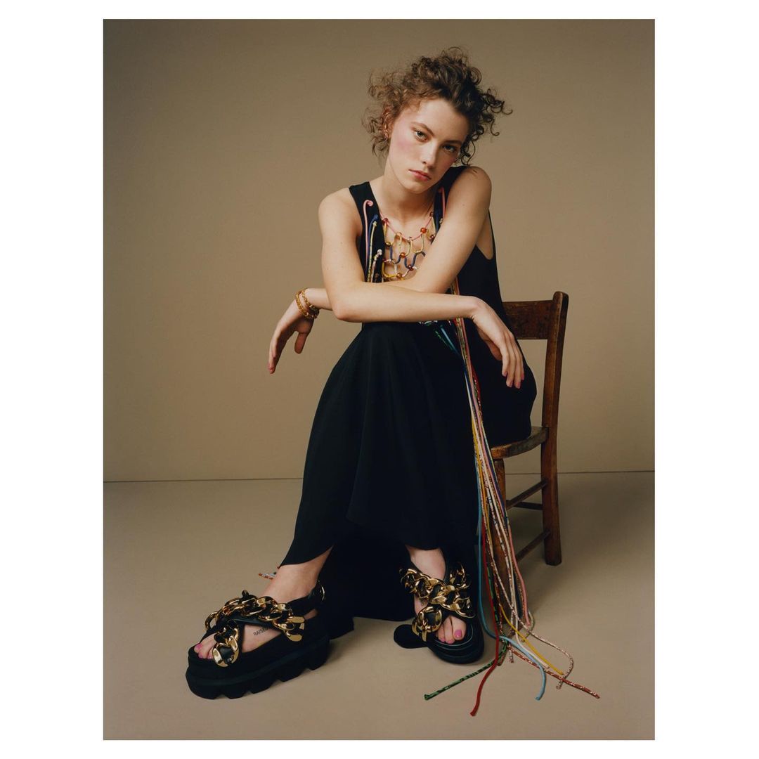 A woman with curly hair sits on a chair wearing a black dress, chunky sandals, and jewelry against a neutral backdrop in a studio setting.