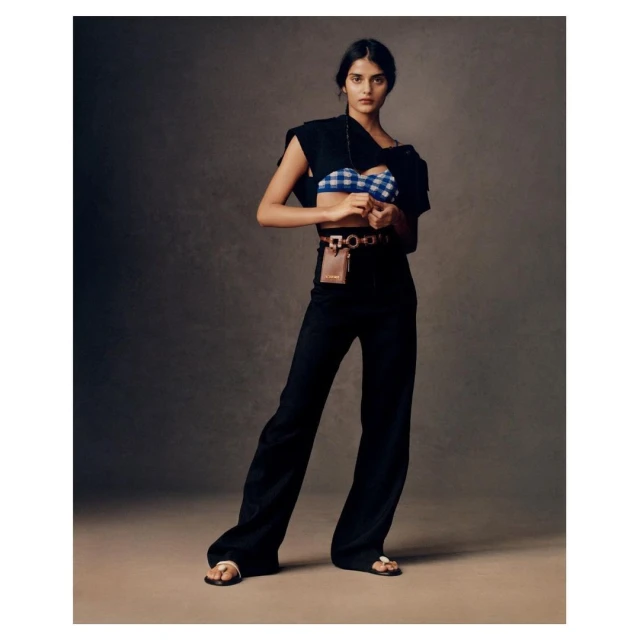 Full-length shot of a woman in a blue and white top, black pants, and sandals, accented by a stylish crossbody bag and belt in studio.