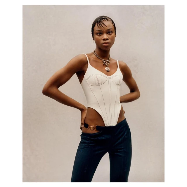 An adult female model poses in a white corset top and dark blue pants against a neutral background, hands on hips, looking directly at the camera.
