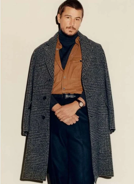 Portrait of an adult male with a mustache wearing a wool plaid overcoat, brown button-down shirt, and black pants posing indoors.