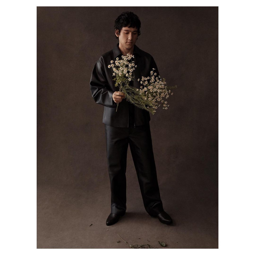 Young man in a dark leather jacket and pants stands holding a bouquet of white daisies against a neutral brown background.