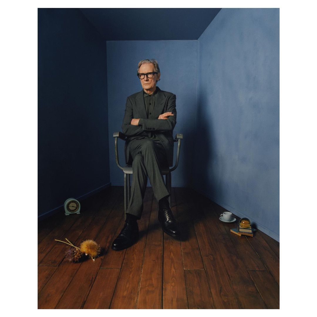 Portrait of an older adult male with glasses, in a grey suit, sitting on a chair in a blue room with a wood floor.