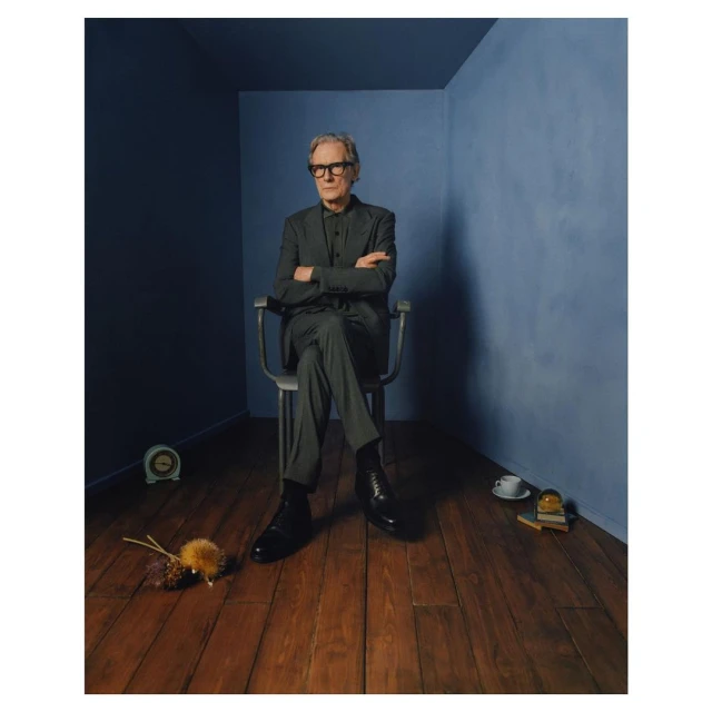 Portrait of an older adult male with glasses, in a grey suit, sitting on a chair in a blue room with a wood floor.
