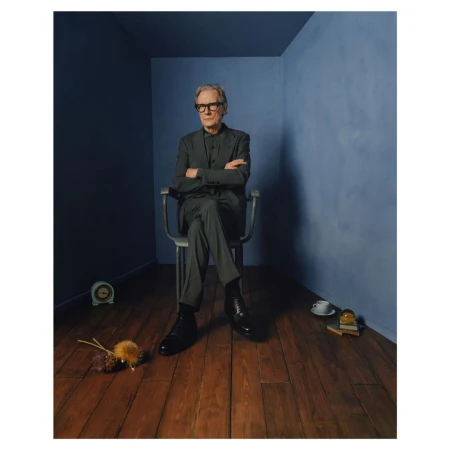 Portrait of an older adult male with glasses, in a grey suit, sitting on a chair in a blue room with a wood floor.