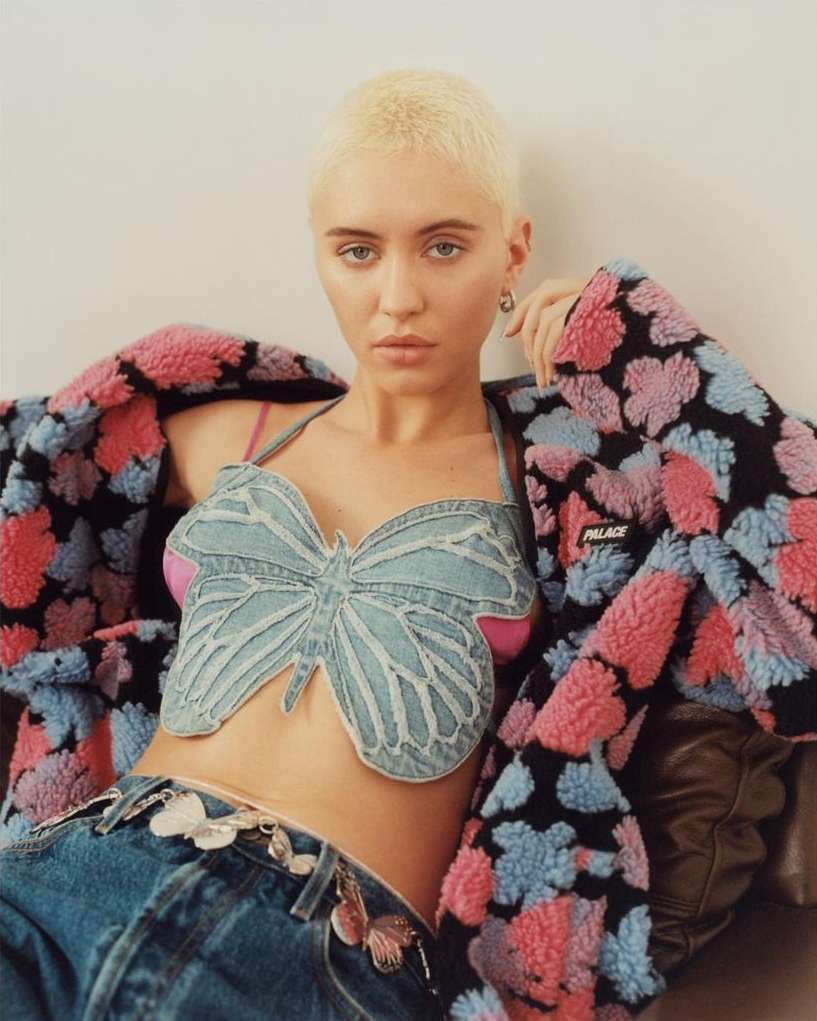 A young adult woman with short blonde hair poses indoors wearing a butterfly denim top, jeans, and a colorful floral jacket.