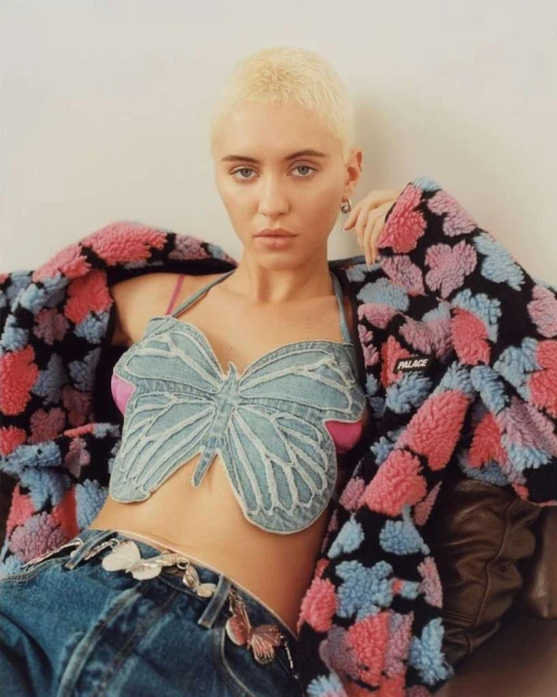 A young adult woman with short blonde hair poses indoors wearing a butterfly denim top, jeans, and a colorful floral jacket.