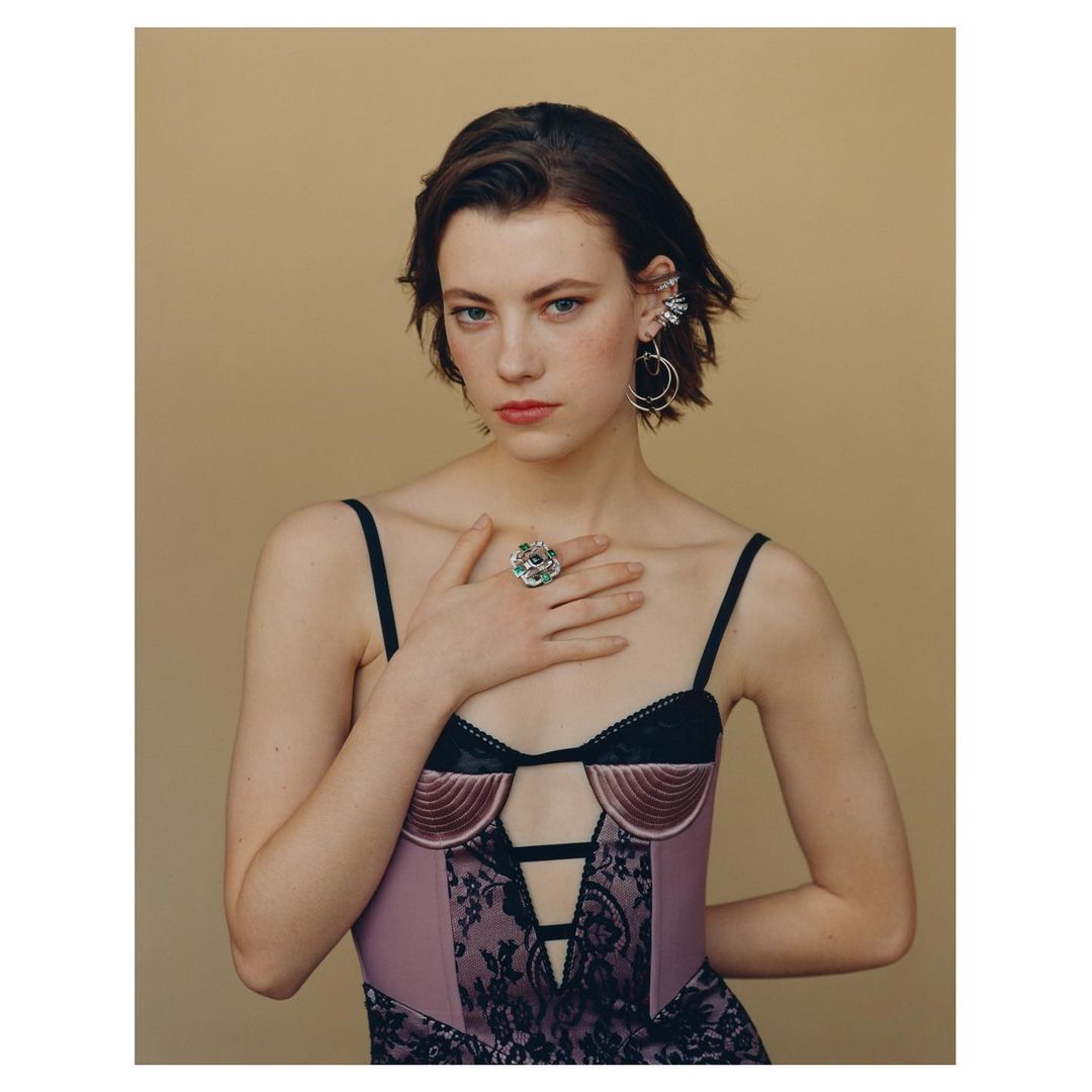 Portrait of a young woman with hand on chest, wearing a lilac bustier with black lace, statement ring and earrings on a neutral background.
