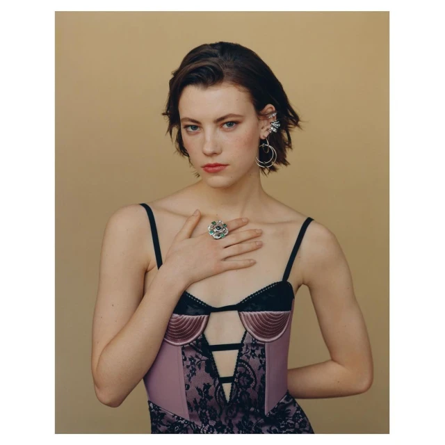 Portrait of a young woman with hand on chest, wearing a lilac bustier with black lace, statement ring and earrings on a neutral background.