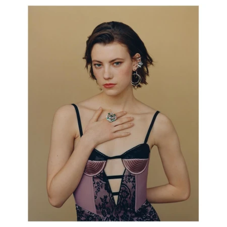 Portrait of a young woman with hand on chest, wearing a lilac bustier with black lace, statement ring and earrings on a neutral background.
