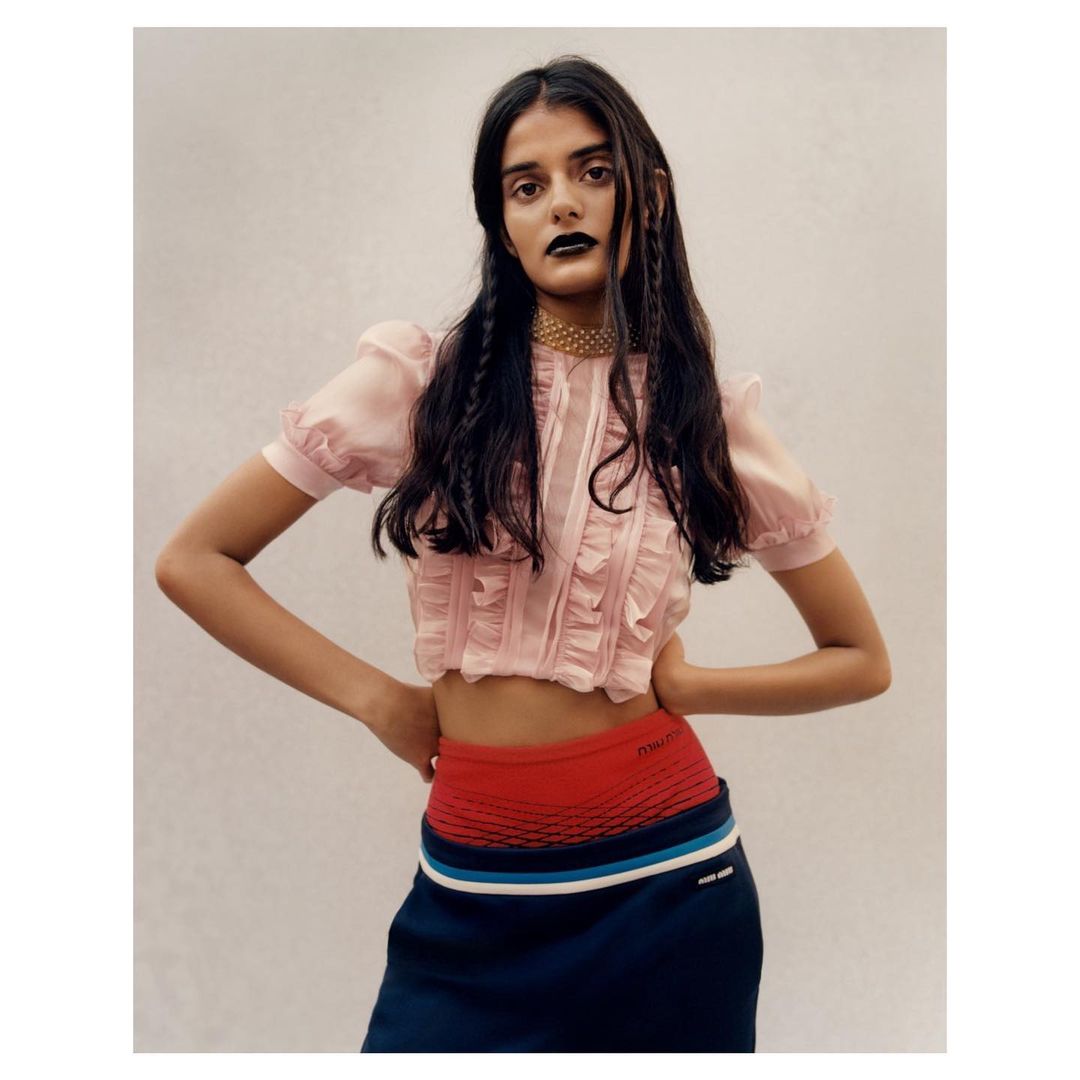 Young woman in a light pink crop top with ruffles and dark blue skirt, posing indoors with hands on her hips, fashion portrait.