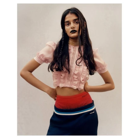 Young woman in a light pink crop top with ruffles and dark blue skirt, posing indoors with hands on her hips, fashion portrait.