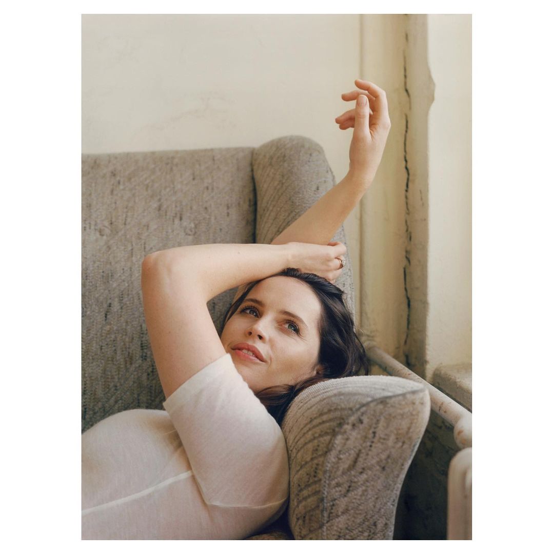 Adult woman lounges in a grey armchair wearing a white top, with her arm raised. The setting is an interior space with a neutral color palette.
