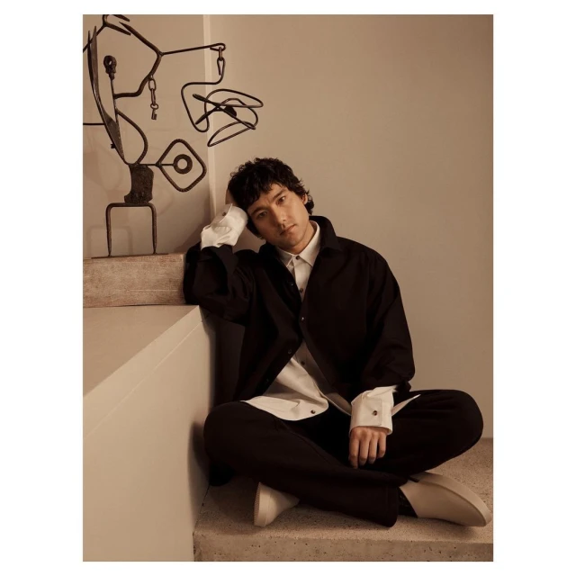 Portrait of a seated adult male wearing a black button-down shirt over a white shirt, positioned near abstract metal art on a staircase.