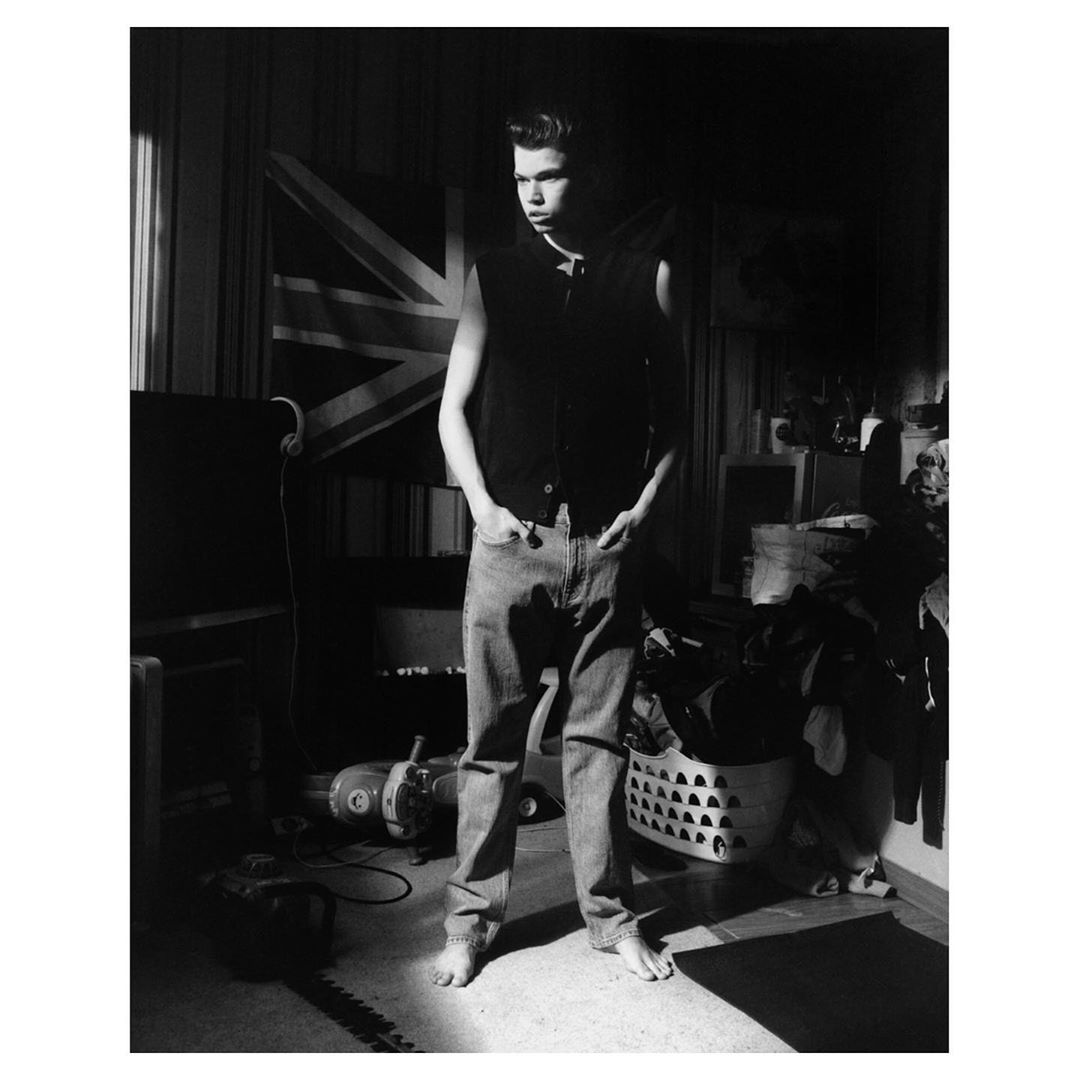 A young adult stands in a room with a Union Jack flag, wearing jeans and a sleeveless shirt, captured in a black and white medium shot.