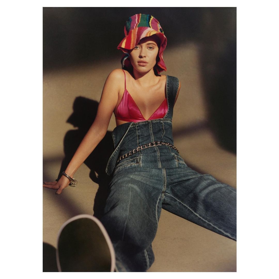 A model in a pink top and denim overalls is seated indoors with a skateboard beside her, wearing a colorful hat, captured in soft lighting.