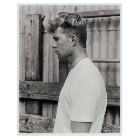 Portrait of a young white man in profile wearing a white t-shirt against a textured wooden fence background.