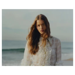 A portrait of a young woman with long brown hair wearing a white ruffled blouse outdoors by the ocean shore.