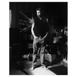 Monochrome portrait of a young man standing in a dimly lit room with hands in pockets wearing a dark vest and jeans