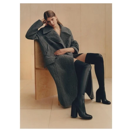 Model wearing textured grey coat and black over-the-knee leather boots sitting on a wooden chair against a plain backdrop.