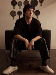 Man with dark hair seated in a dark leather armchair wearing a black t-shirt and light slip-on shoes against a wall with paddles