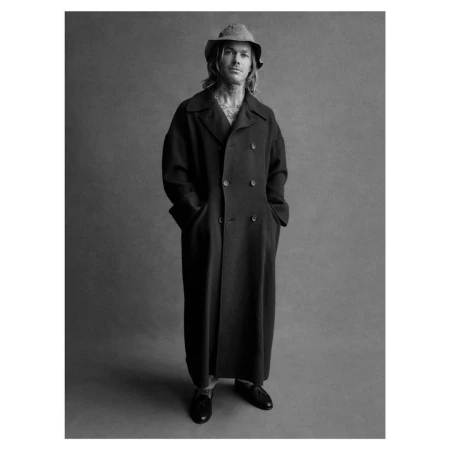 Man wearing a long dark coat and bucket hat standing against a textured grey studio backdrop