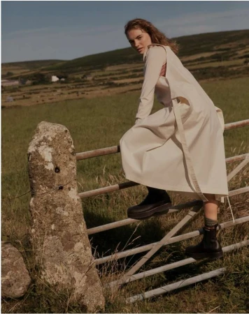 Woman in cream dress and dark boots climbing a metal farm gate outdoors in a grassy rural landscape