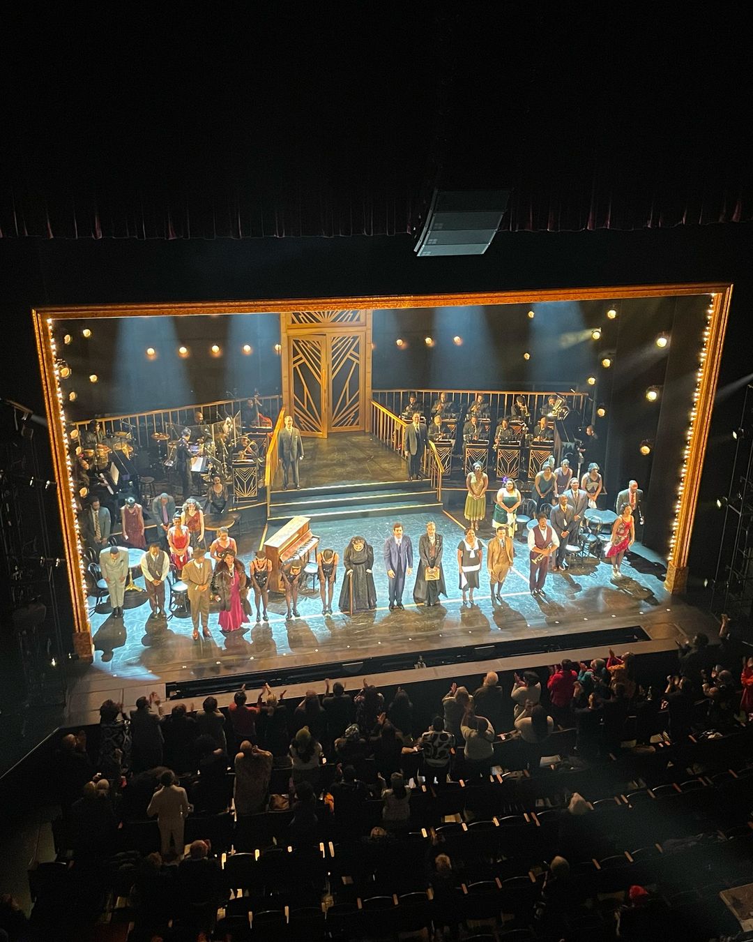 Theatre stage with actors, musicians, and audience. People on stage are bowing to audience. Gold-trimmed stage framing detail.