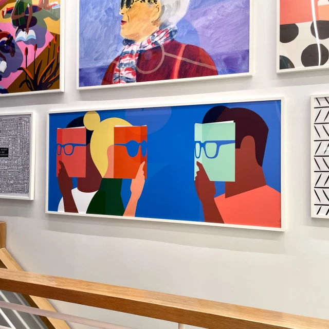 Art print featuring stylized figures reading books in front of their faces, displayed among other framed artworks on a white wall.
