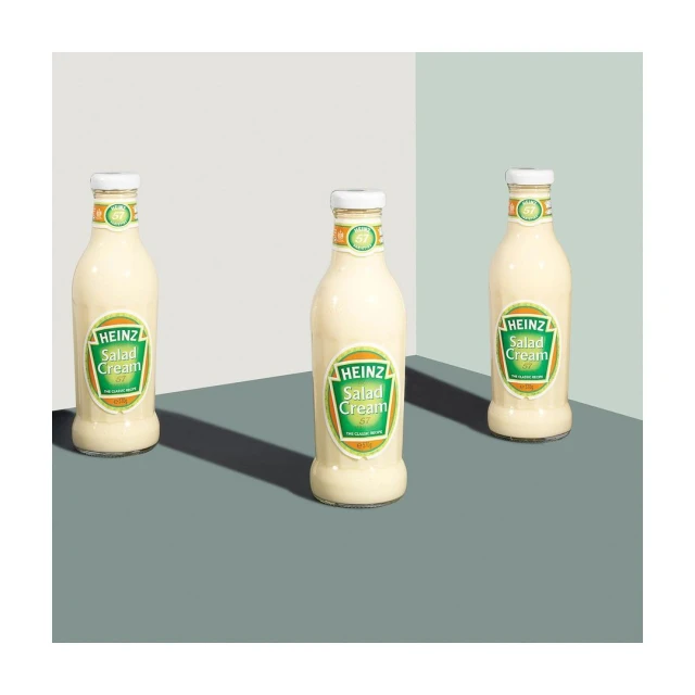 Three bottles of Heinz Salad Cream stand on a grey surface against a two-tone grey and white background in a product shot.