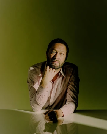 Portrait of a man with short dark hair and a beard wearing a striped shirt, posed against a green backdrop.