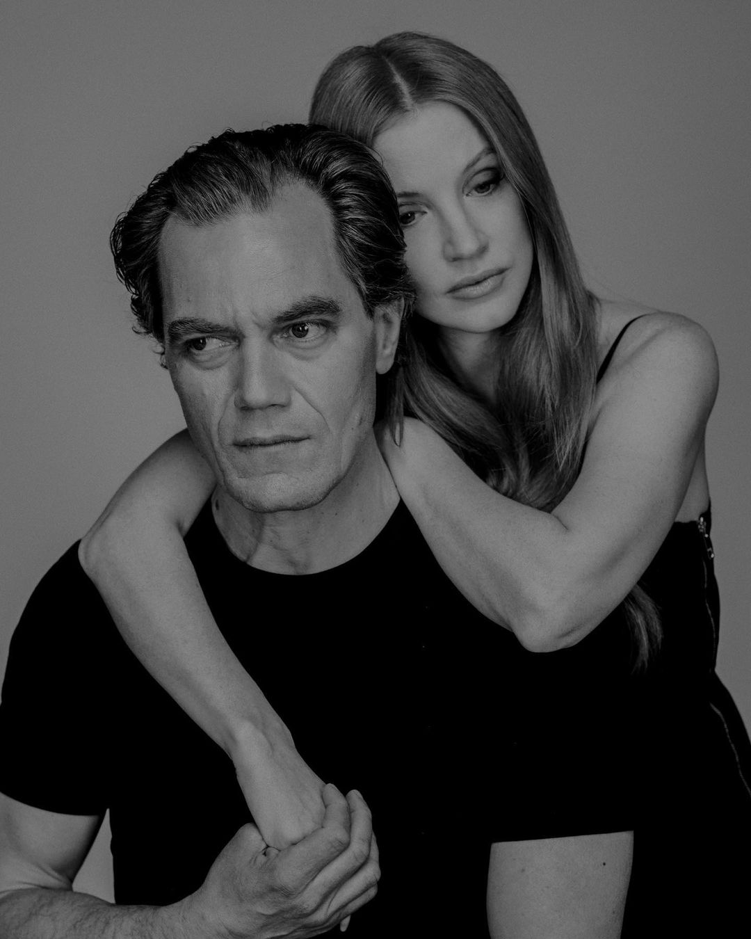 Black and white portrait of a man and woman in dark clothing. The woman stands behind the man with her arms over his shoulders.