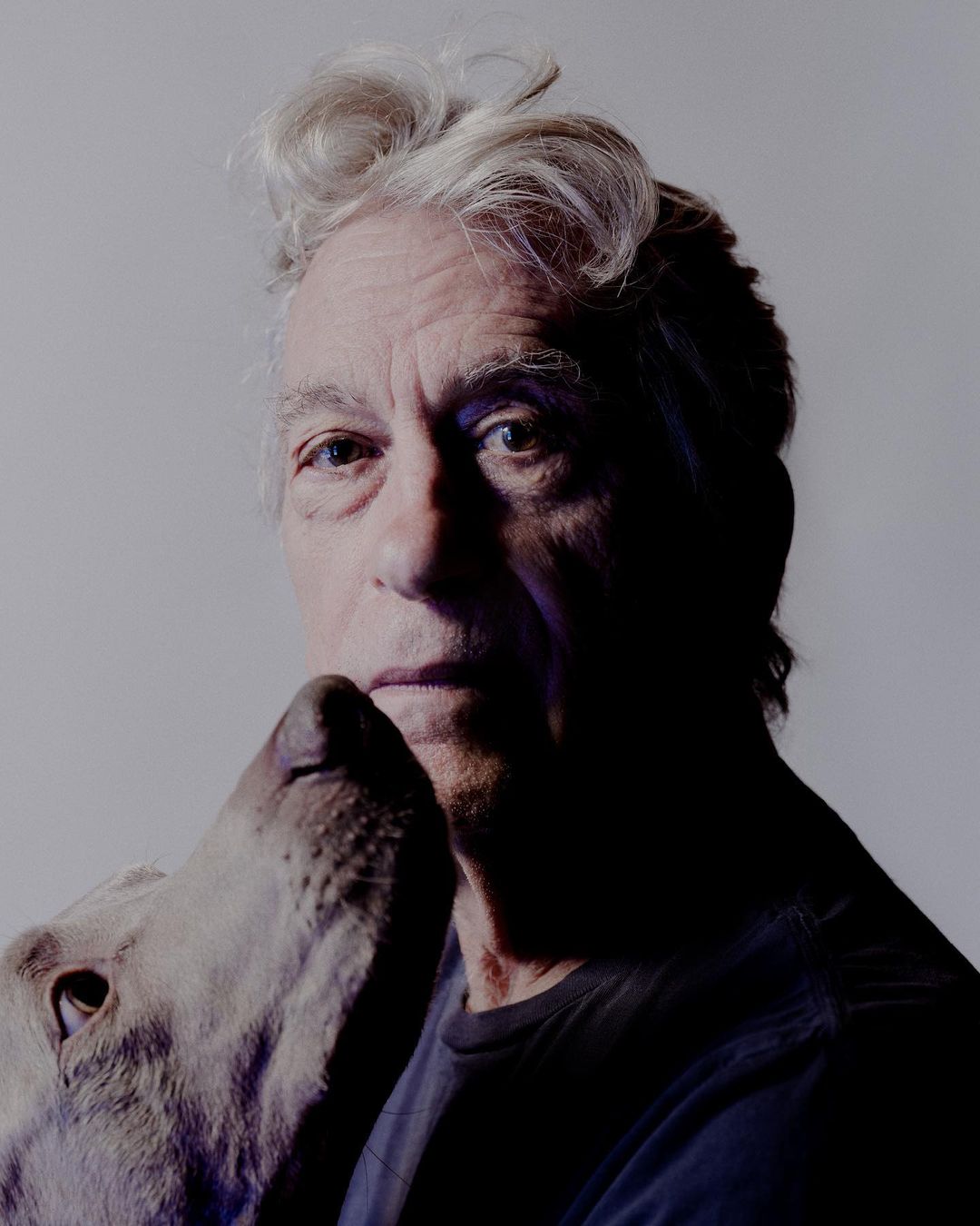 Close-up portrait of an elderly man with a gray dog's head near his face, set against a gradient gray studio backdrop with soft lighting.