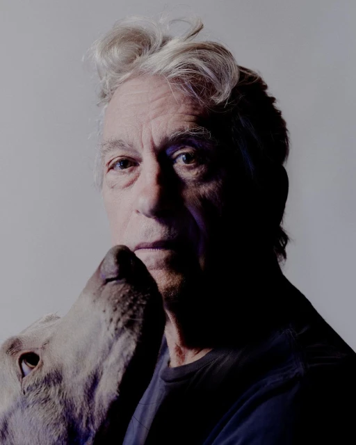 Close-up portrait of an elderly man with a gray dog's head near his face, set against a gradient gray studio backdrop with soft lighting.