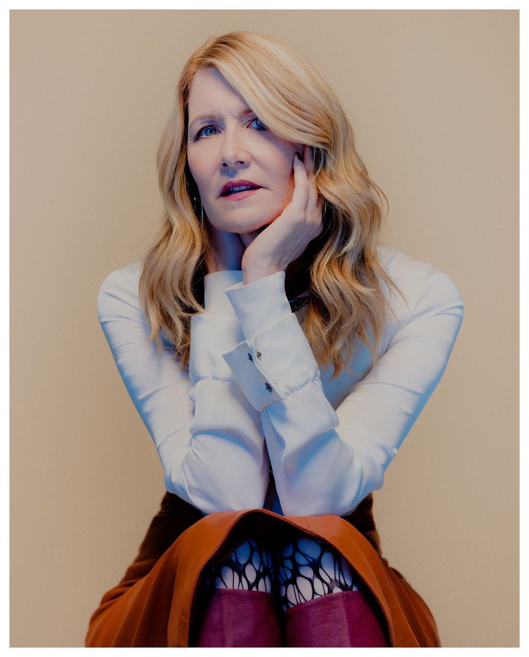 Portrait of fair-skinned adult woman with blonde wavy hair, wearing a white top, rust-colored skirt, and black tights against a neutral background.