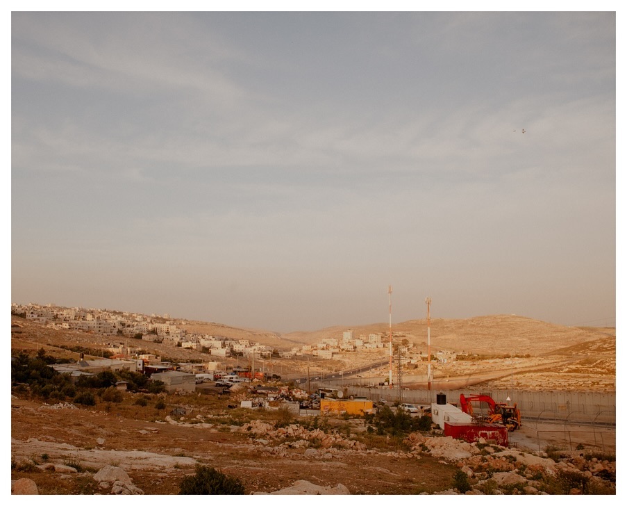 Panoramic view of a desert landscape featuring small settlements, hills, a winding road, and rugged terrain under a pale sky during daytime.