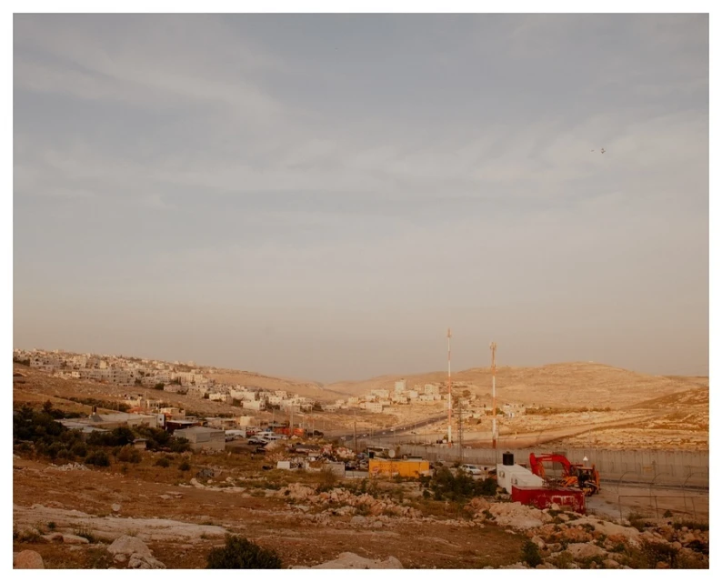 Panoramic view of a desert landscape featuring small settlements, hills, a winding road, and rugged terrain under a pale sky during daytime.