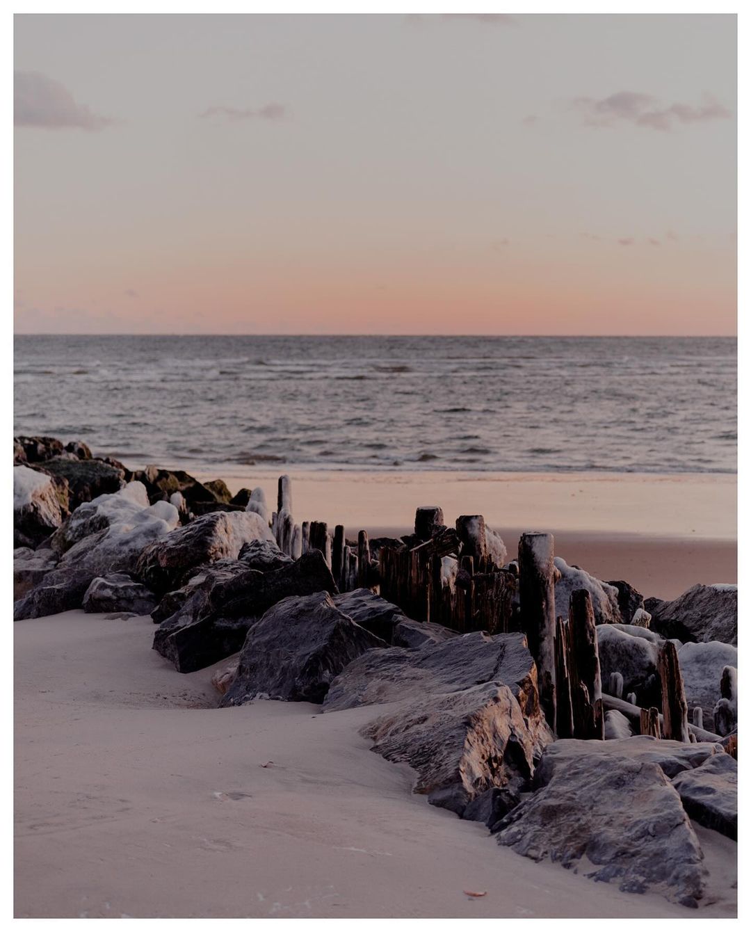 Seascape photograph with waves meeting sand on the beach at sunset. Rocks and wooden pilings line the shoreline in the foreground.