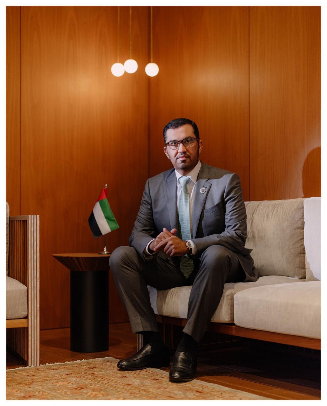 Portrait of an adult male in a grey suit sitting on a couch indoors in front of a wood paneled wall next to an Emirati flag.