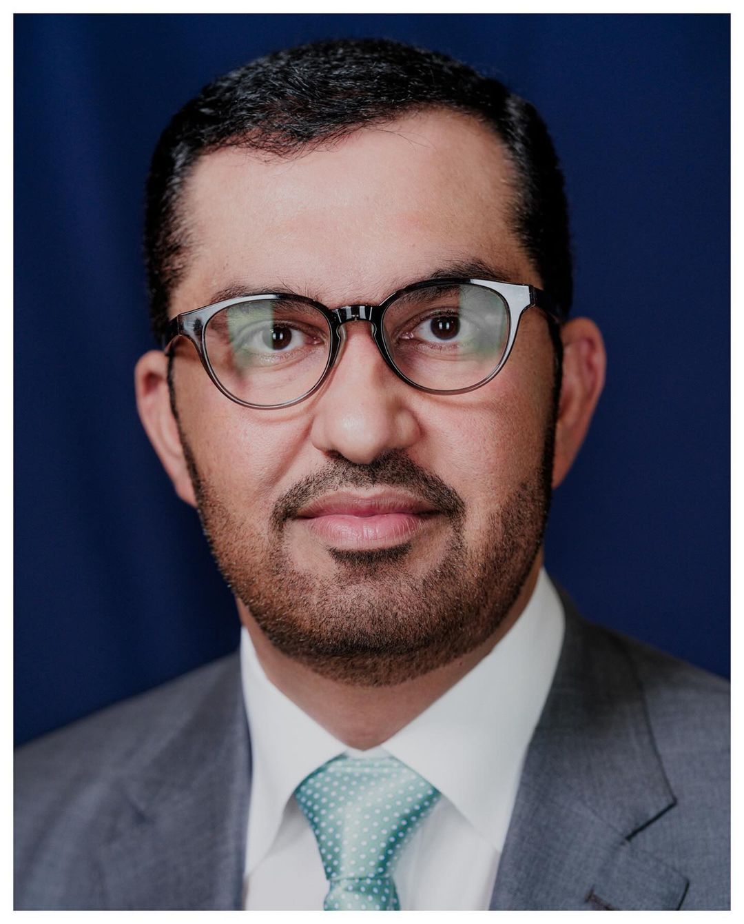 Headshot of a middle-aged man wearing glasses, a suit, and a patterned tie, set against a dark blue backdrop, with a focus on the face.
