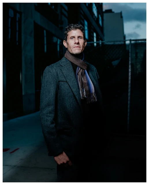 Adult man in gray overcoat and striped scarf stands in an urban outdoor setting, captured in medium shot under dim lighting conditions.