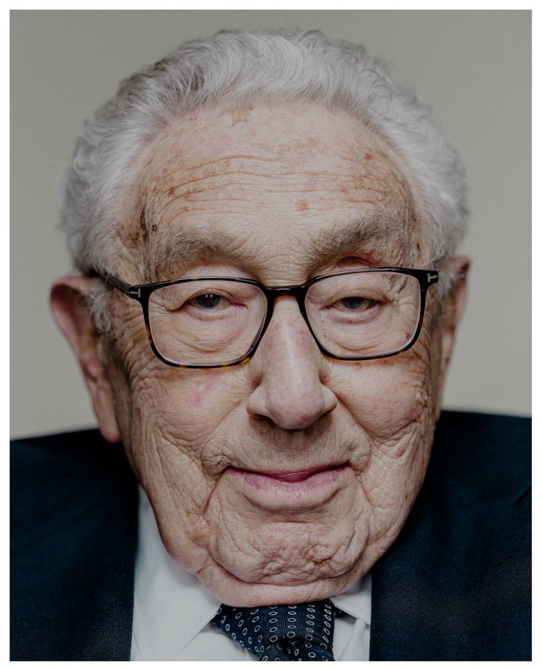Close-up portrait of an elderly man with gray hair wearing glasses, a dark suit, and a patterned tie against a neutral background.