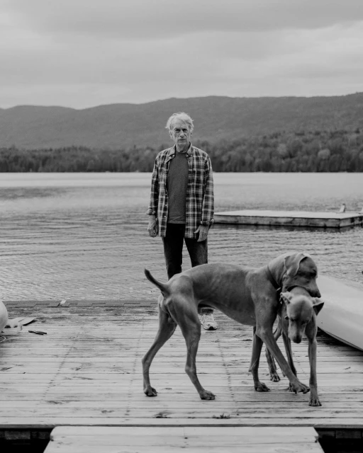 Black and white image of an adult man standing by a lake with two Weimaraner dogs on a wooden dock with forested mountains behind them.