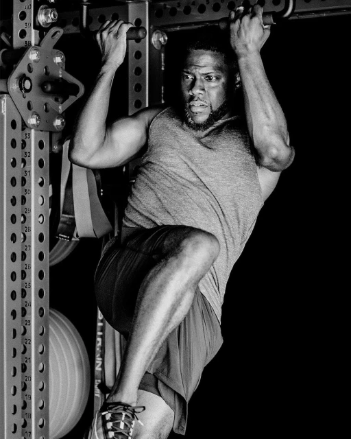 Black and white image of Kevin Hart doing pull-ups. Hart is in workout clothes and gym equipment is visible in the background.