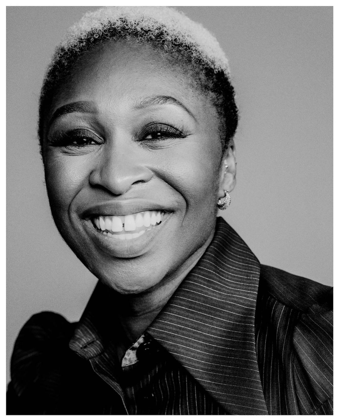 Close-up portrait of an adult Black woman with short white hair smiling broadly, wearing a dark pinstripe collared shirt against grey.